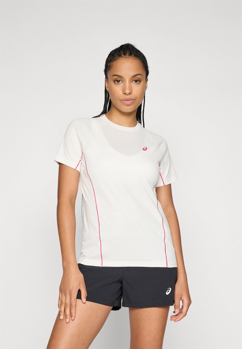 Kvinde med fletninger iført en hvid sports t-shirt med pink detaljer og sorte shorts, stående foran en ensfarvet lys baggrund.