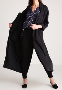 Mustat pitkä takki, jossa on rento malli, leveät hihat ja etunapit, kerrostettuna tummansinisen paidan kanssa, jossa on valkoisia tähtikuvioita. Mustat housut ja ballerinat.