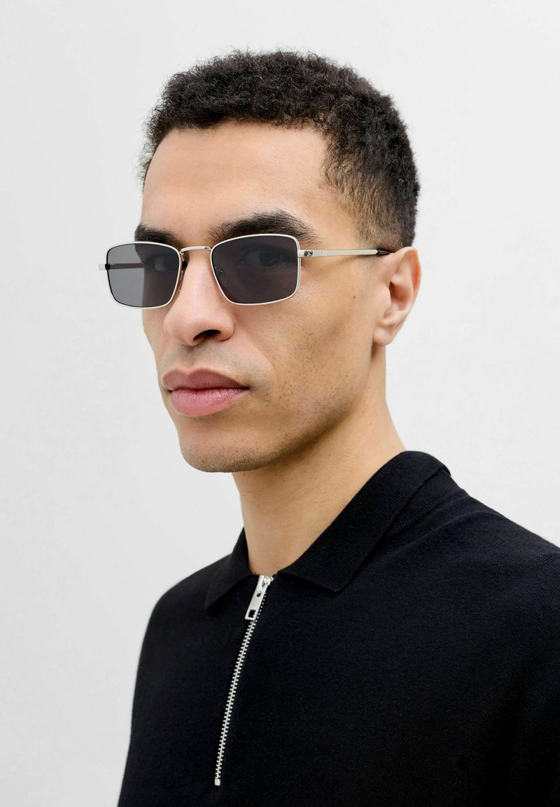 Homme aux cheveux courts et bouclés portant des lunettes de soleil carrées noires et une chemise noire à col zippée, sur un fond clair uni.