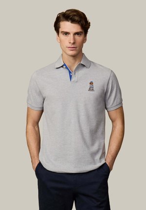 Joven con camisa tipo polo gris claro con pequeño logo bordado de oso y pantalones azul marino, de pie con las manos en los bolsillos frente a un fondo liso.