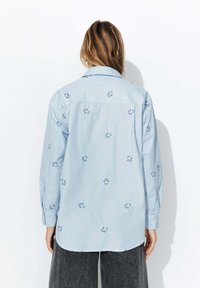 Chemise rayée bleu clair avec broderie florale dans des tons de bleu, manches longues, col et ourlet arrondi.