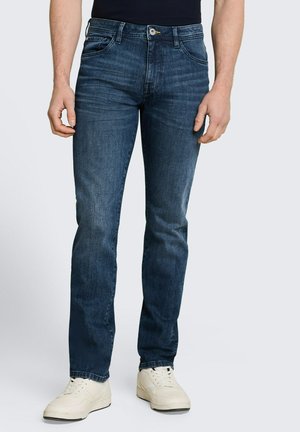 Mężczyzna ubrany w ciemnoniebieskie jeansy slim-fit i białe sneakersy stoi na prostym jasnym tle.