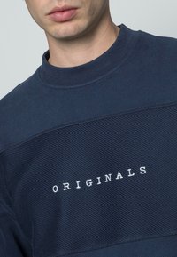 Felpa navy con collo a giro, caratterizzata da un tessuto testurizzato e colletto a coste, decorata con la scritta bianca "ORIGINALS" sul petto.