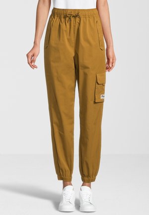 Vrouw draagt mosterdkleurige cargo joggerbroek met elastische taille en manchetten, gecombineerd met witte sneakers.