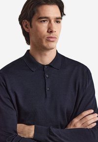 Marinblå långärmad pikétröja med platt krage, fyrknappsstängning och ribbade ärmslut, med en slät textur och skräddarsydd design.