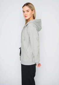 Felpa grigia con cappuccio in materiale morbido, dotata di cappuccio regolabile, tasche laterali e un taglio rilassato. Accenti neri nella parte inferiore.