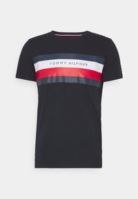 Černé tričko s krátkým rukávem, které má vodorovný proužkový design v námořnické modré, bílé a červené. Na přední straně je vytištěné logo "TOMMY HILFIGER".