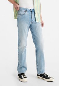 Personne portant un jean bleu clair, des baskets noires et blanches, une chemise blanche et une chemise à rayures jaunes partiellement visible sur fond blanc.