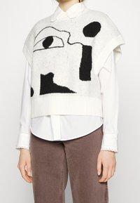 Weißer Strick-Sweater-Weste mit schwarzen abstrakten Mustern, breiten Armausschnitten und geripptem Saum. Getragen über einem weißen Hemd mit Kragen.