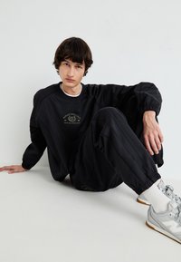 Hanorac negru și pantalonii asortați cu manșete elastice, având text și logo aurii. Purtați cu adidași gri și șosete albe.