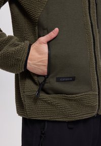 Hand, die in eine Reißverschlusstasche einer olivgrünen Fleecejacke mit strukturiertem Stoff und "ICEPEAK"-Etikett greift.