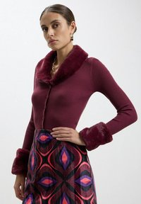 Cardigan bordeaux a coste con colletto e polsini in pelliccia morbida, abbinato a una gonna decorata con forme geometriche in rosa e viola.