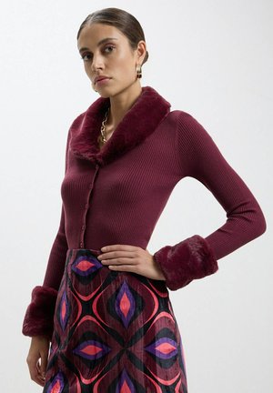Cardigan bordeaux a coste con colletto e polsini in pelliccia morbida, abbinato a una gonna decorata con forme geometriche in rosa e viola.