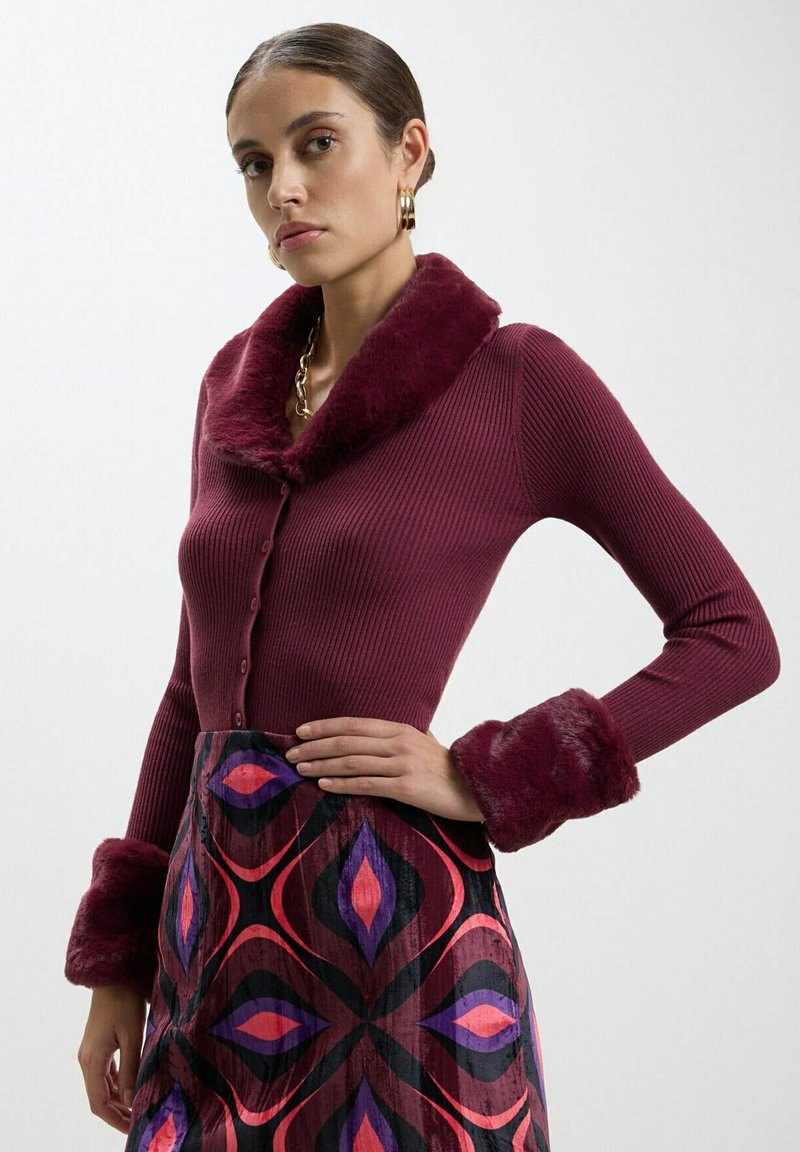 Cardigan bordeaux a coste con colletto e polsini in pelliccia morbida, abbinato a una gonna decorata con forme geometriche in rosa e viola.