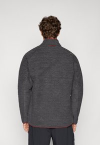 Szürke polárdzseki magas gallérral, texturált felülettel és piros varrással az alján. Raglánujjakkal rendelkezik és egyszerű dizájnja van.