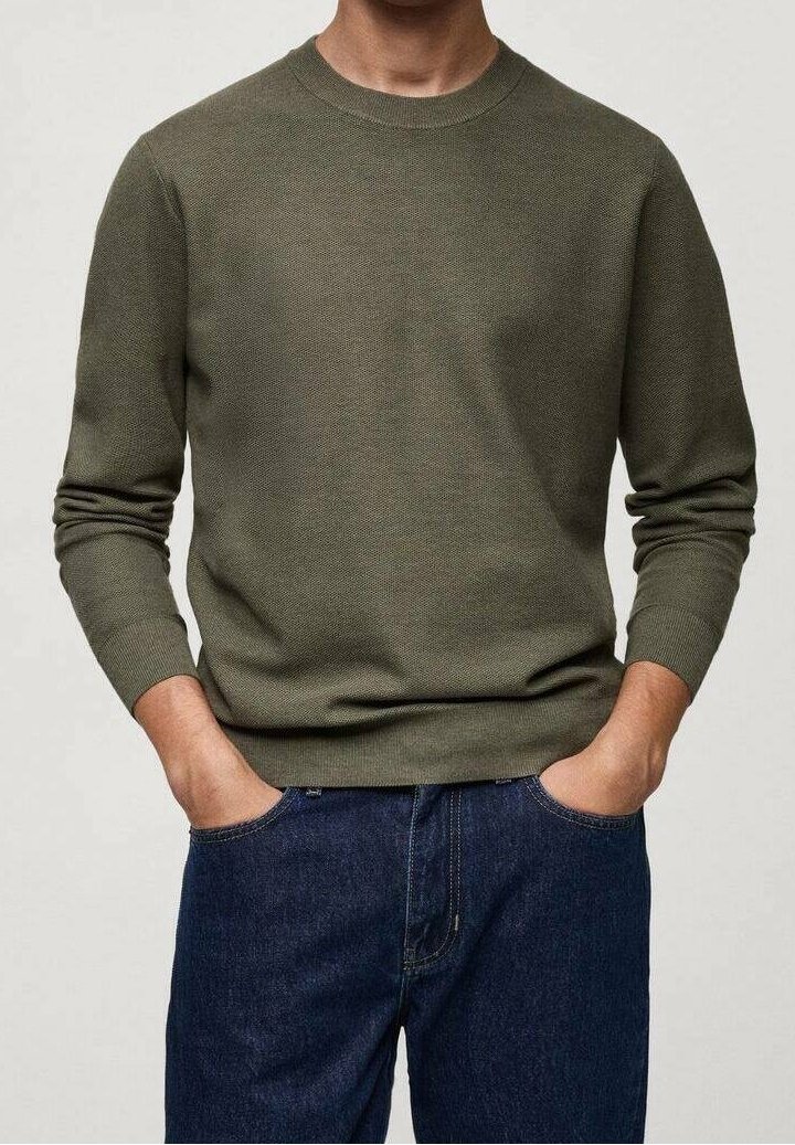 Homme portant un pull vert olive à manches longues et un jean bleu foncé, les mains dans les poches, sur un fond uni.