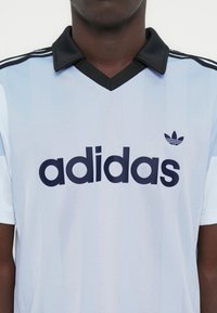 Ανοιχτό μπλε ποδοσφαιρική φανέλα με μαύρες λεπτομέρειες, που διαθέτει μαύρο γιακά, τρεις μαύρες ρίγες στους ώμους και ένα μεγάλο σκούρο λογότυπο στο εμπρός μέρος.