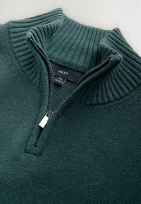 Donkergroene gebreide trui van medium dikte met geribde hoge kraag en gedeeltelijk openstaande halve ritssluiting, waarbij het maat- en landlabel binnenin zichtbaar is.