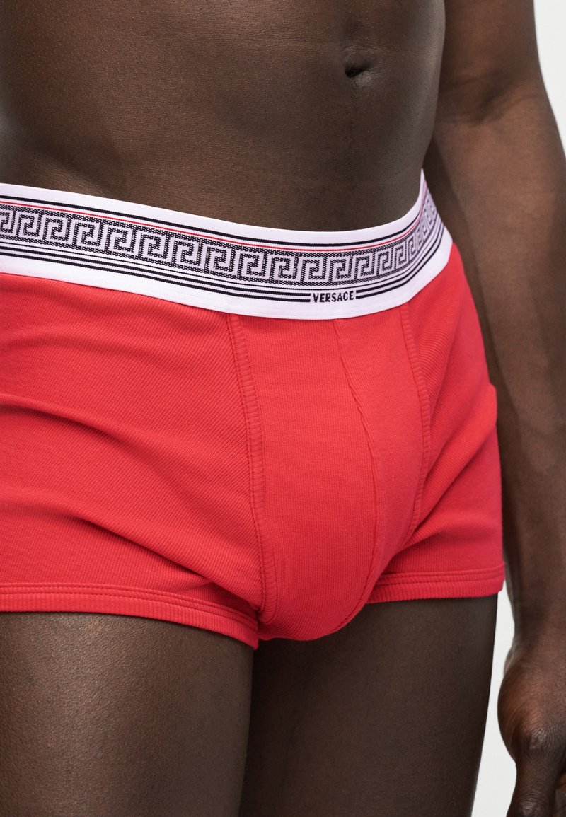 Mannelijk torso gekleed in rode boxershorts met een witte tailleband, voorzien van een zwart geometrisch patroon en het Versace-logo aan de voorkant.