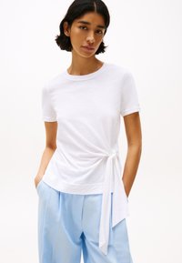 Tricou alb cu mânecă scurtă, realizat dintr-un material ușor, are un detaliu legat la nivelul taliei și este asortat cu pantaloni de un albastru deschis.