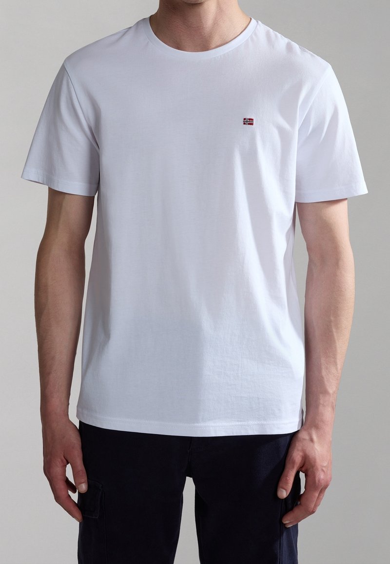 White cotton t-shirt with a crew neck, featuring a small red and black flag emblem on the left chest. Smooth texture, relaxed fit.