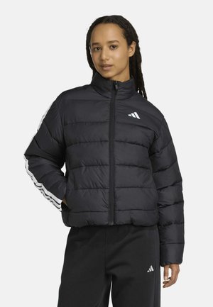 Mujer con chaqueta acolchada negra con rayas blancas en las mangas y pantalones negros, de pie con una mano en el bolsillo de la chaqueta.