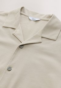 Beige kortärmad skjorta med en mjuk textur, utrustad med krage, tre knappar och sydda detaljer.