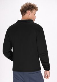 Zwarte longsleeve-shirt met een zachte textuur, platte kraag en een subtiele naaddetail op de achterkant. Gdraag met blauwe broek.