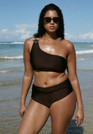 Mujer de cabello rizado con bikini negro de un solo hombro y gafas de sol, parada waist-deep en el agua del océano bajo un cielo azul despejado.