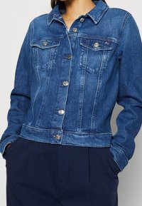 Giacca di jeans in una tonalità blu medio, con due tasche sul petto anteriori, bottoni argentati e un design classico del colletto. Tessuto testurizzato.