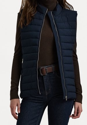 Kvinde iført marineblå quiltet ærmeløs vest over brun rullekrave, mørke jeans og brunt bælte med sølvspænde.