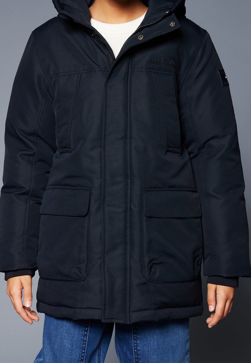 Personne portant un manteau d'hiver noir à capuche avec des poches avant et un pull blanc en dessous, associé à un jean bleu.