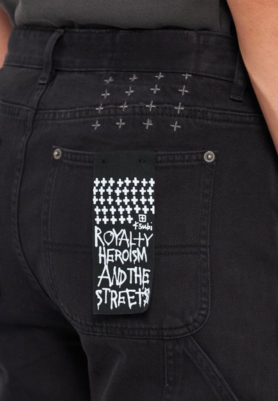 Black denim jeans featuring a stitched design of small crosses on the back pocket and a tag with white graphic text and black background.