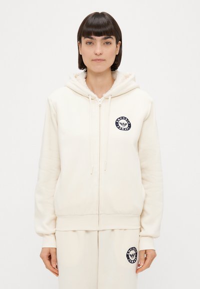 Cremfarbener Zip-Hoodie mit einem fleecegefütterten Kapuzen, gerippten Bündchen und Saum. Ausgestattet mit einem kreisförmigen Marineblau-Logo auf der Brust. Weiche Stofftextur.