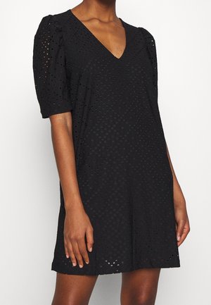 Vrouw draagt een zwarte jurk met korte mouwen van eyelet-stof en een V-hals, staand tegen een effen witte achtergrond.