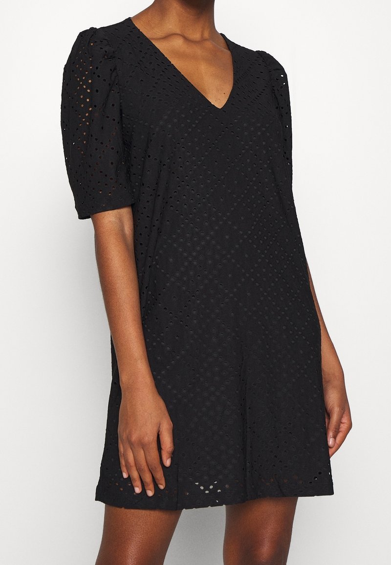 Femme portant une robe noire à manches courtes en tissu ajouré avec un décolleté en V, debout devant un fond blanc uni.