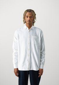 Camisa blanca de manga larga con botones en un tejido texturizado, que incluye un bolsillo en el pecho y un corte relajado. Llevada con pantalones de mezclilla oscuros.
