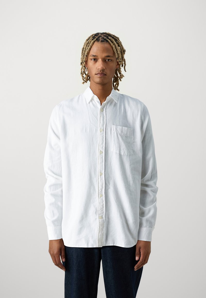 Camisa blanca de manga larga con botones en un tejido texturizado, que incluye un bolsillo en el pecho y un corte relajado. Llevada con pantalones de mezclilla oscuros.
