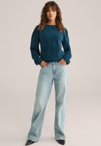 Teal texturerad tröja med långa ärmar, rund halsringning, tillsammans med ljusblå högmidjade jeans och svarta spetsiga skor.
