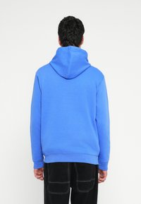 Modra jopa s kapuco iz mehke tkanine, z velikim prostorom za kapuco. Ima rebraste manšete in spodnji rob, združena s črnimi cargo hlačami. Enostaven, neokrašen dizajn.