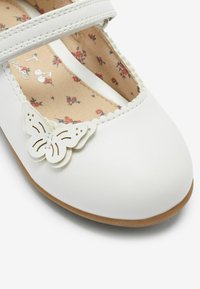Vita läder Mary Jane-sko med en ensam rem, scalloped kant och en fjäril som accent. Inredningen har blommig tryck. Slät textur.