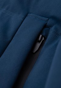 Gros plan d'un tissu bleu marine avec une fermeture éclair noire partiellement visible sous un rabat en tissu.