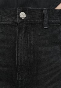 Melnai džinsiem ir redzams sudraba logotipa pogas, smalka tekstūra un standarta piecu kabatu dizains ar kontrastējošiem šuvju elementiem.