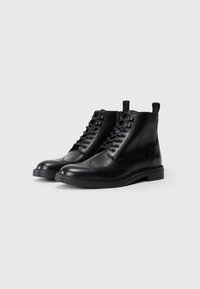 Botine negre din piele, cu sireturi, cu detalii brogue și mânere pentru o mai bună fixare, pe fundal alb.
