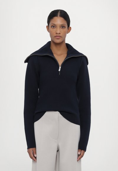 Navy blue zip-up sweater with a high collar and long sleeves. Fabric appears soft with a smooth texture. Worn over light beige trousers.