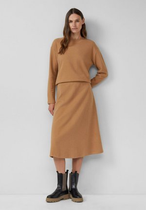 Frau steht vor einer grauen Wand und trägt einen camelfarbenen Pullover sowie einen Midirock mit schwarzen Stiefeletten.