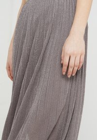 Gonna plissettata di un grigio metallico con un tessuto morbido e testurizzato, che mostra linee verticali e un leggero luccichio. Mano che riposa delicatamente sul tessuto.