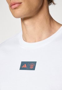 Hvitt T-skjorte med rund hals har et rektangulært grafikk i mørkeblått og korall, som viser Adidas-logoen og Bayern München-emblemet.