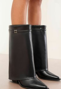 Fekete bőrből készült bokacsizma, magas, strukturált szárakkal. Behajtott felsőrésszel és fémes logóval díszítve, valamint hegyes orrú kialakítással.