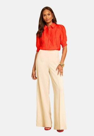 Femme portant un chemisier rouge vif avec des manches bouffantes et un pantalon large crème taille haute ainsi que des sandales rouges, posant avec un bracelet en or.